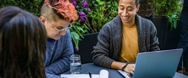 man in orange crew neck shirt using laptop beside two people