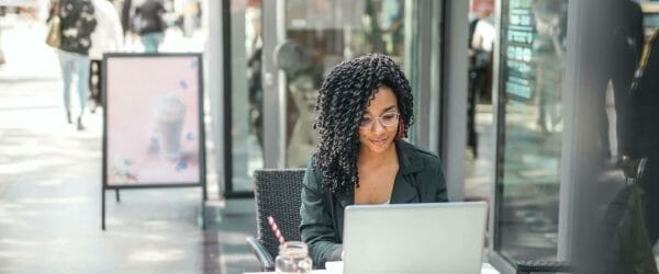 ethnic young woman using laptop while having tasty beverage in modern street cafe