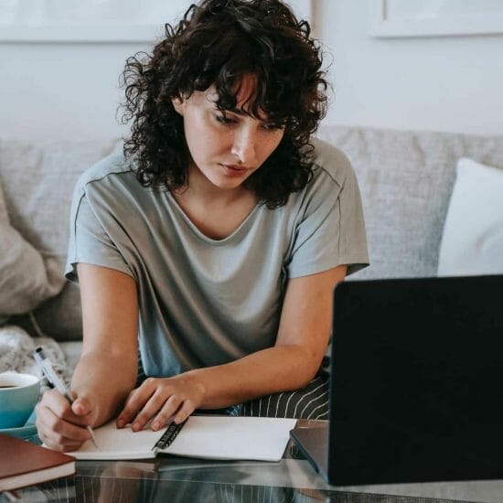 serious young lady writing in notebook during online studies on laptop at home