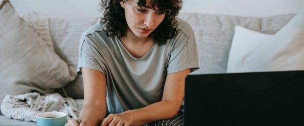 serious young lady writing in notebook during online studies on laptop at home