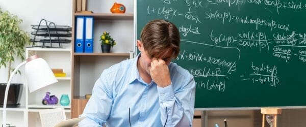 young male teacher wearing glasses tired overworked touching his nose closed eyes sitting school desk with books notes front blackboard classroom