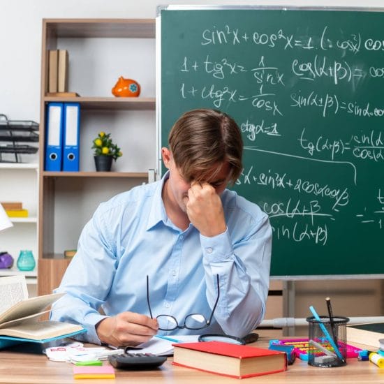 young male teacher wearing glasses tired overworked touching his nose closed eyes sitting school desk with books notes front blackboard classroom