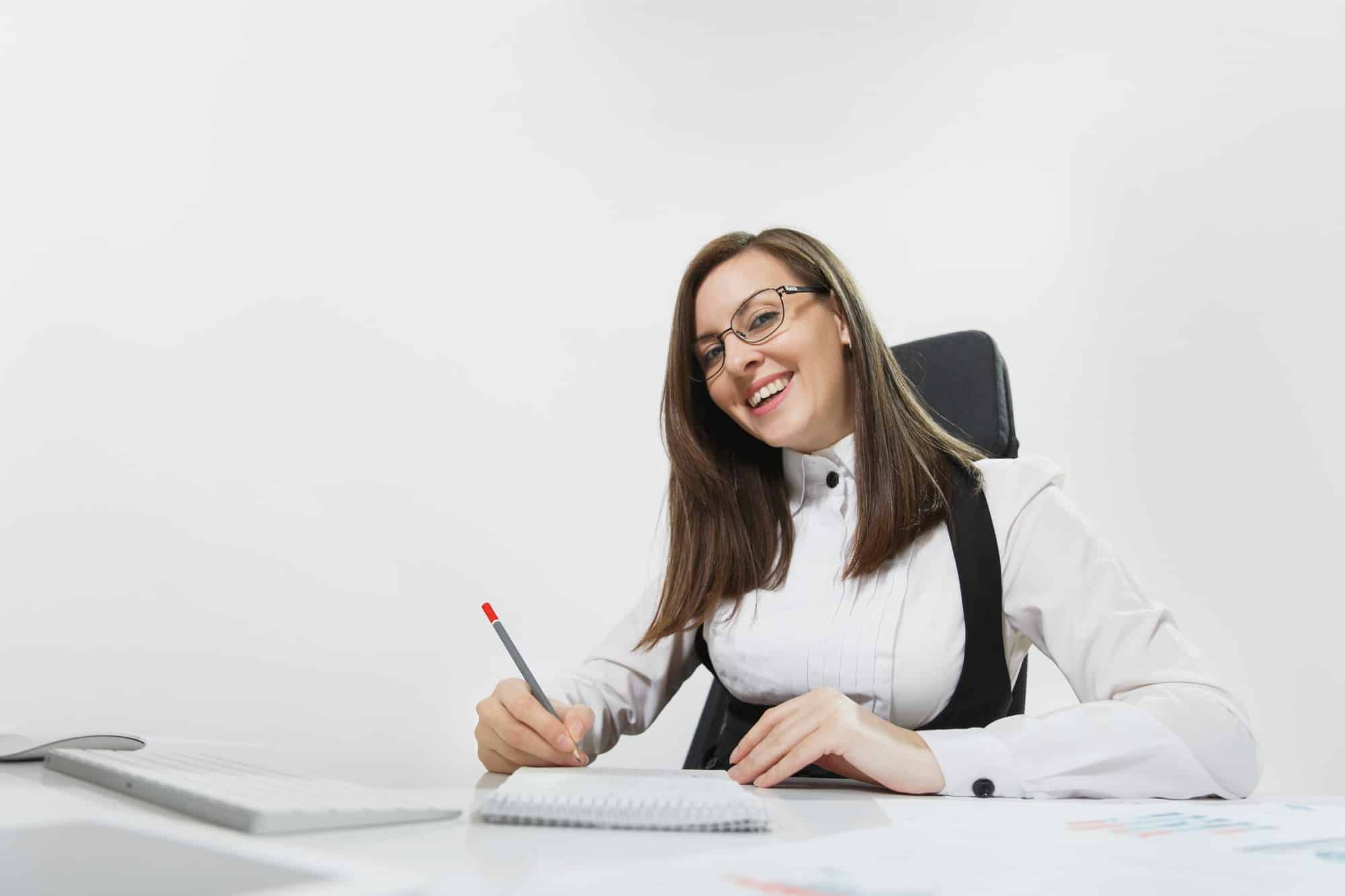 beautiful smiling brown hair business woman suit glasses sitting desk working computer with modern monitor documents light office writing notebook