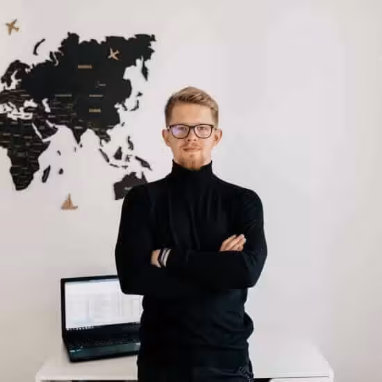 indoor-portrait-handsome-blond-man-wearing-spectaculars-black-pullover-posing-white-wall-with-world-map-laptop-desktop