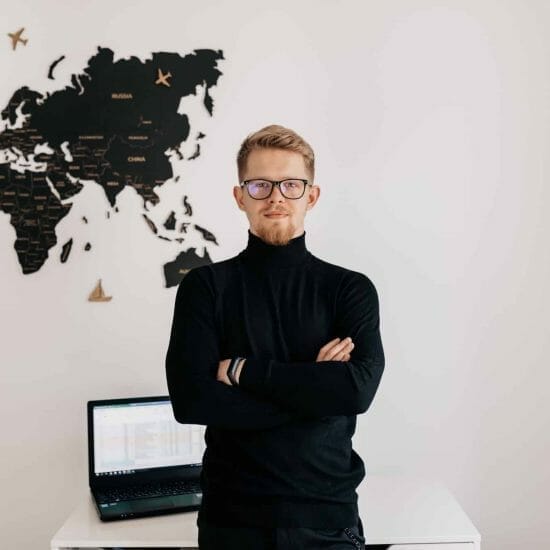 indoor-portrait-handsome-blond-man-wearing-spectaculars-black-pullover-posing-white-wall-with-world-map-laptop-desktop