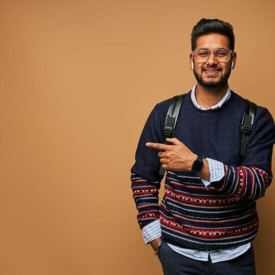 smiling happy indian student with backpack pointing his finger wall