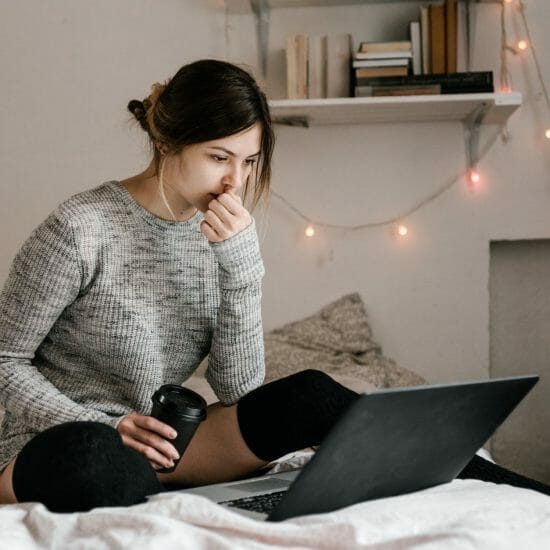 confused woman in gray sweater sitting on bed using macbook