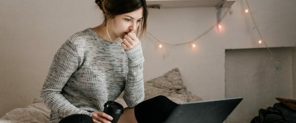 confused woman in gray sweater sitting on bed using macbook